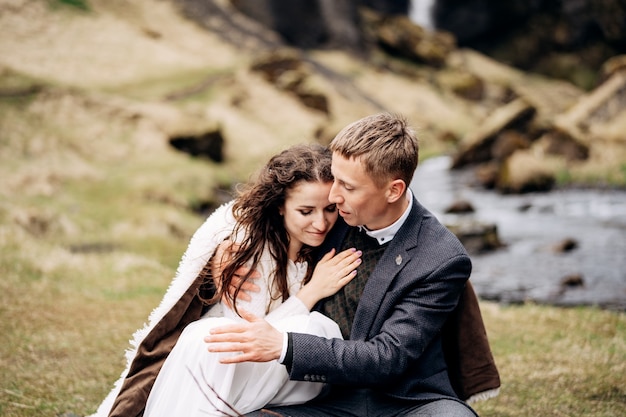 Destination Iceland wedding, near Kvernufoss waterfall. A wedding couple sits on the banks of a