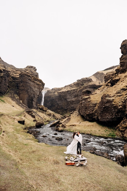 Destination Iceland wedding, near Kvernufoss waterfall. A wedding couple stands under a plaid near a mountain river. The groom hugs bride. They built an impromptu wedding table with decor and guitar