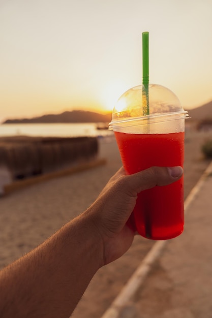 Detail of an unrecognizable man39s hand holding a glass of slushy drink in the foreground at sunset on the beach