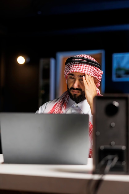 Detailed view of tired man in Arabian attire sitting at office desk and using laptop to conduct internet research. Muslim man with minicomputer on the table, looking weary and suffering from headache.