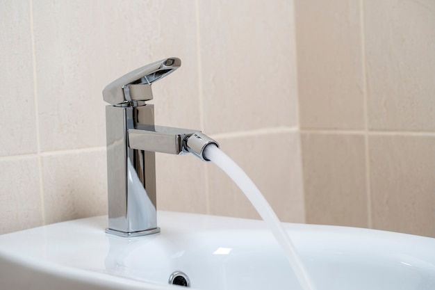 Details of white ceramic bidet with water running from tap in modern bathroom with beige tiles