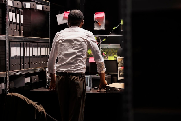 Detective analyzing crime weapon and photos attached to wall, searching insight. African american police investigator looking at evidence and photographs connection map at board