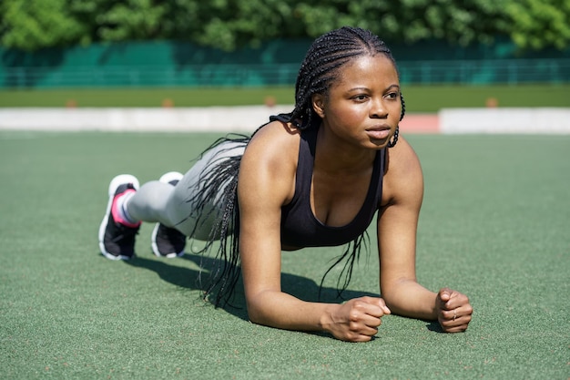 Determinated black woman does plank exercise on green turf flooring of city stadium against park