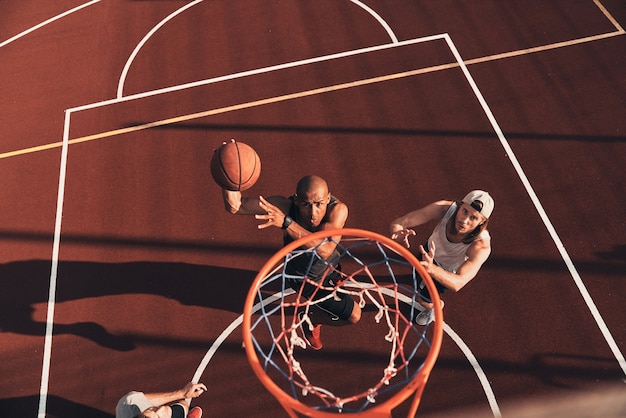 Determined to win. Top view of young man in sports clothing scoring a slam dunk while playing basketball with friends outdoors