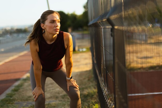 Determined woman rests against a fence after an intense workout session during the golden hour at an outdoor track and field facility