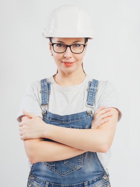 A determined woman in a white hard hat and denim overalls stands with her arms crossed exuding confidence and professionalism in construction