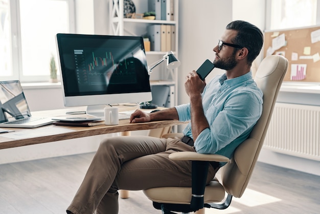 Developing new approaches. Young modern businessman using smart phone and smiling while sitting in the office