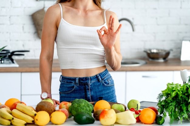 Diet Concept. Portrait of a healthy young woman with nice figure near the table with fruits and vegetables showing ok gesture, concept of healthy food.