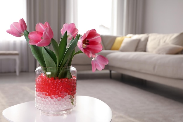 Different color fillers and tulips in glass vase on white table at home space for text Water beads