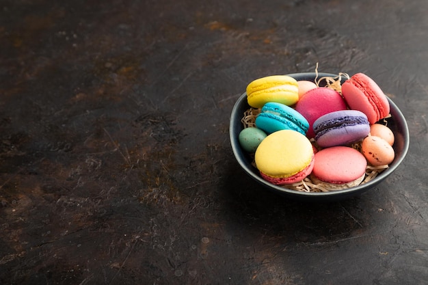 Different colors macaroons and chocolate eggs in ceramic bowl on black concrete background side view copy space