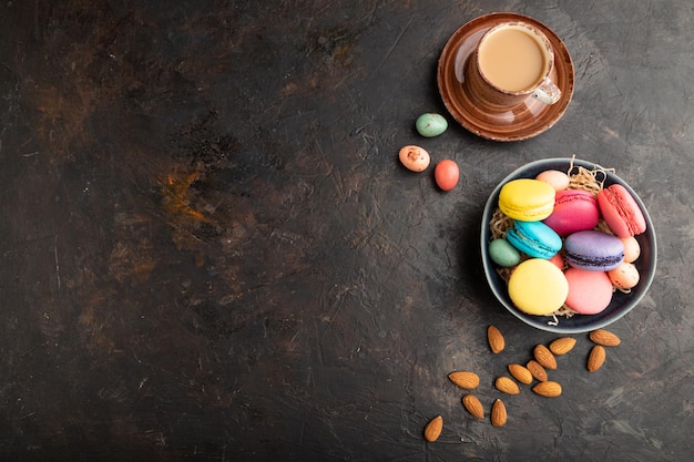 Different colors macaroons and chocolate eggs in ceramic bowl cup of coffee on black concrete background top view copy space