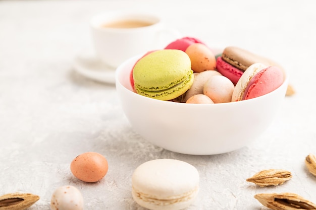 Different colors macaroons and chocolate eggs in ceramic bowl cup of coffee on gray concrete background side view close up