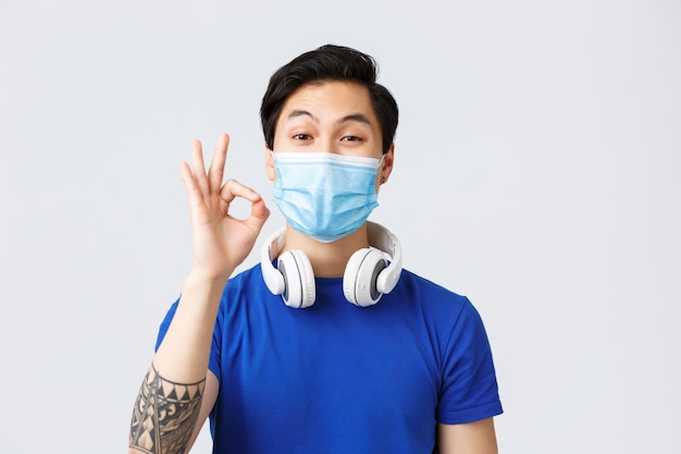 Different emotions, lifestyle and leisure during coronavirus, covid-19 concept. Pleased good-looking asian guy in medical mask take-off headphones to show okay sign, likes music or song