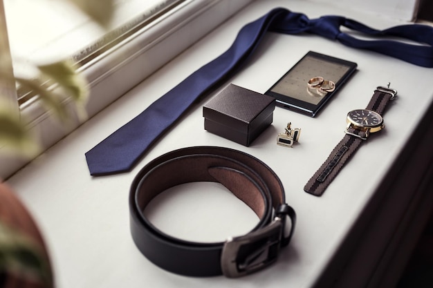 Different men's accessories such as cufflinks watches rings belt tie and phone are on the table