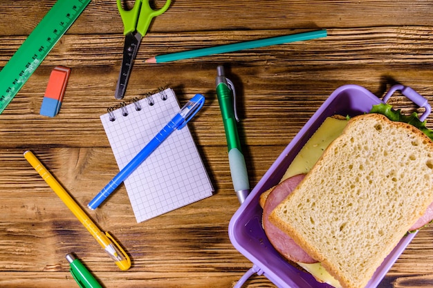 Different stationeries and lunch box with sandwiches on a wooden table Top view