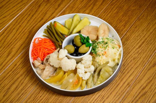 Different types of pickled vegetables on a white plate. Pickled cucumbers, tomato cherry, greens, mushrooms and garlic with onion and lettuce on the table