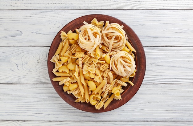 Different types and shapes of Italian pasta in ceramic plate on white wooden background