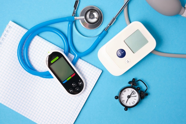 Digital blood pressure monitor, glucometer, stethoscope, notepad and alarm clock on blue background