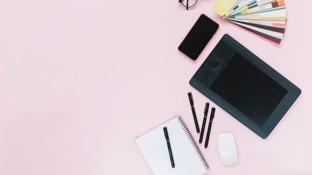 Digital graphic tablet; cellphone and mouse with spiral notebook and felt-tip pens on pink background