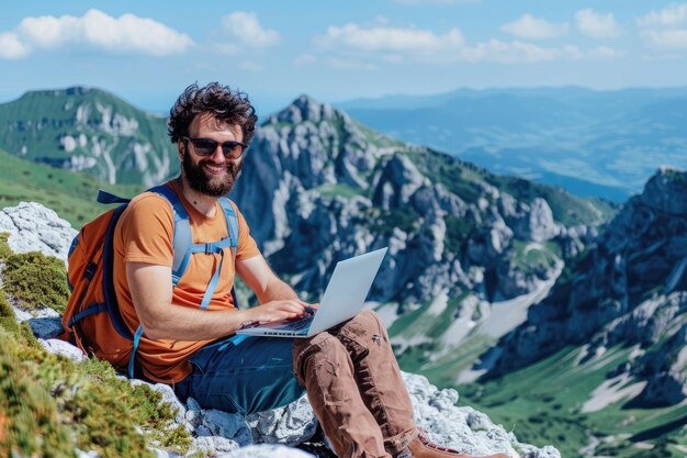 A digital nomad smiling while working on his laptop seated on a mountain peak with a breathtaking view of the landscape around