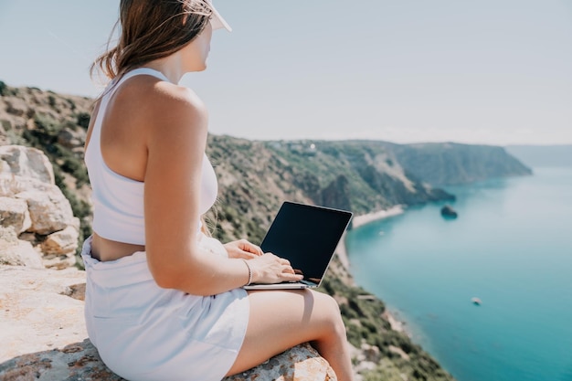 Digital nomad woman in the hat a business woman with a laptop sits on the rocks by the sea during