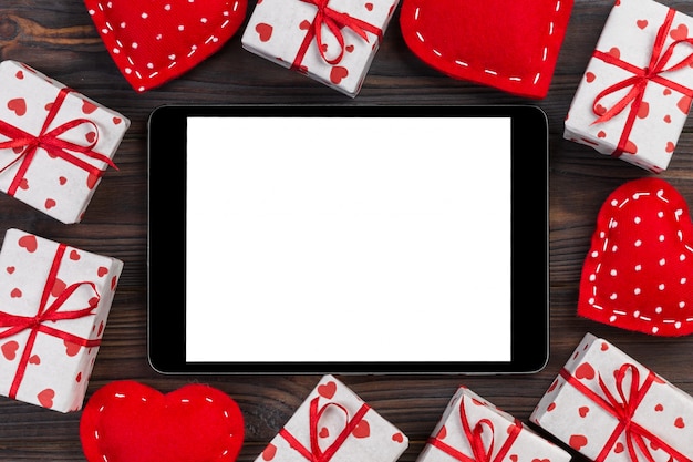 Digital tablet blank screen with gift box and hearts decor on wooden table. Top view.