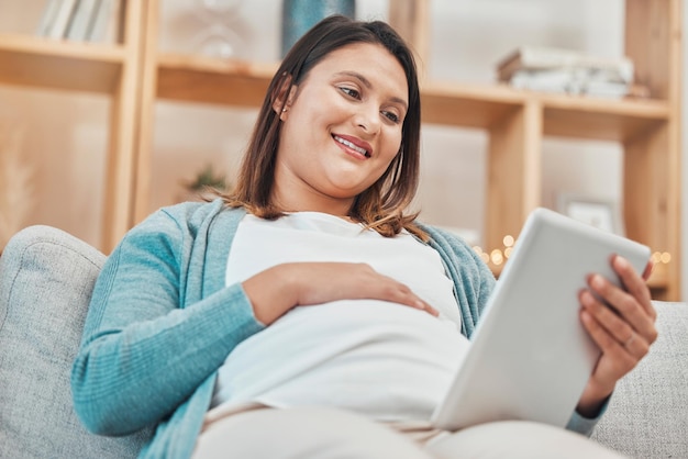 Digital tablet pregnant and happy woman relaxing on a sofa in the living room of her home Calm pregnancy and lady resting and watching a video on social media or a movie online with mobile device