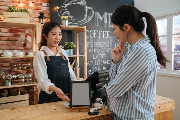 digital tablet with blank screen in coffee shop. coffeehouse female worker hands showing gesture with menu on mobile pad in bar counter and talking to customer. lady client thinking while take order.