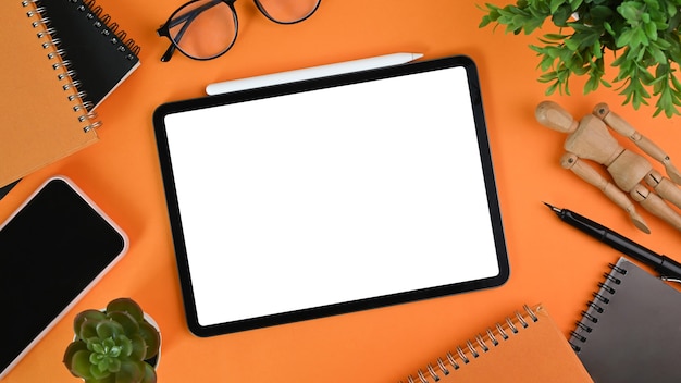 Digital tablet with blank screen, smart phone, house plant ,glasses and notebook orange background.