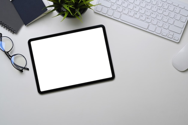 Digital tablet with empty screen, glasses, plant and keyboard on white office desk.