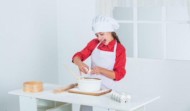 Diligent approach happy child cooking in kitchen bake cookies in kitchen professional and skilled baker kid in chef uniform and hat teen girl preparing dough making cake by recipe time to eat