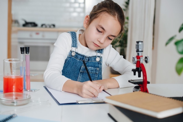 Diligent little girl doing home science project, documenting the process. She has chemistry glassware with colorful liquids and microscope