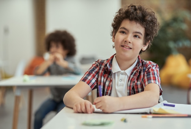 Diligent little school boy listening to his teacher while sitting at the desk in classroom