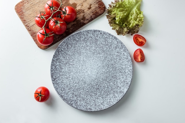 Dinner plate mockup. Gray empty plate in the kitchen with tomatoes and lettuce leaves on a white table. Flat lay, mock up