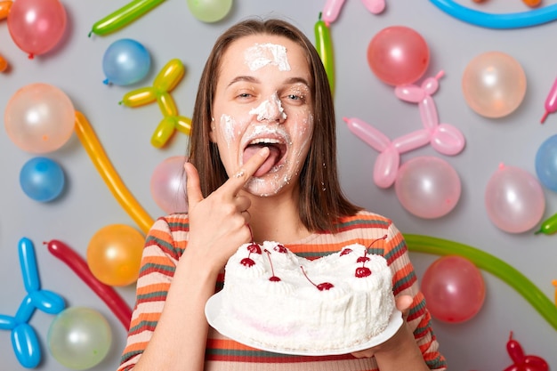 Dirty woman wearing striped dress standing against gray wall with balloons decoration standing with cream on face licking finger eating cake enjoying holiday dessert