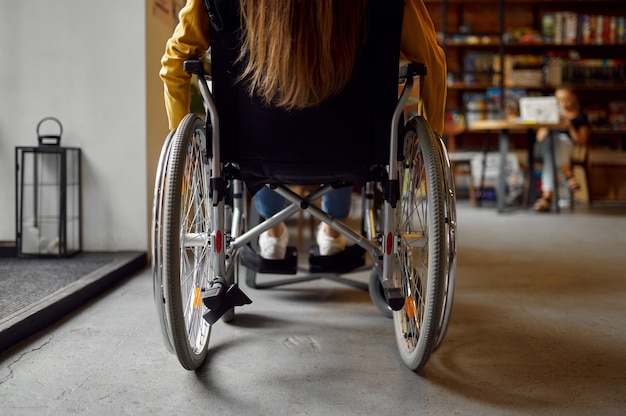 Disabled female student in wheelchair, back view, disability, bookshelf and university library interior on background. Handicapped young woman studying in college, paralyzed people get knowledge