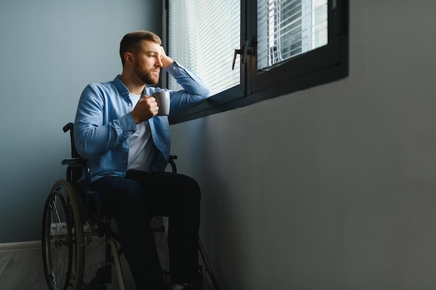 Disabled Person Sits in Wheelchair Against Window Serious Sad Caucasian Man Wearing Casual Clothes and Look at Large Panoramic View in Bright Modern Living Room or Hospital