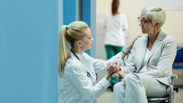 Disabled senior woman in wheelchair holding hands with a doctor while communicating with her in a hallway at clinic