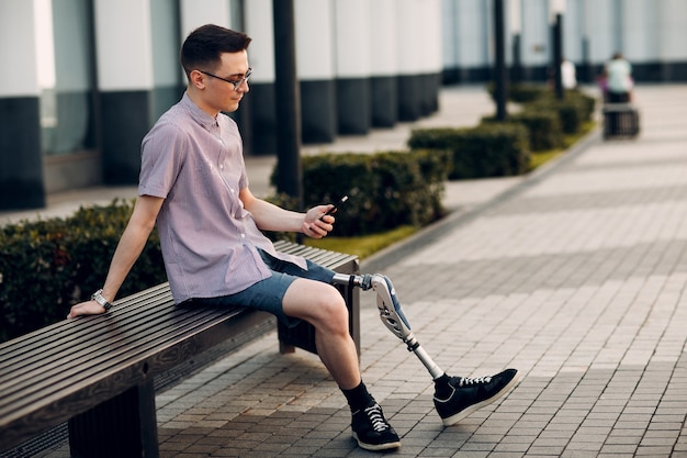 Disabled young man with foot prosthesis sitting on bench on city street and holds mobile phone outdoor