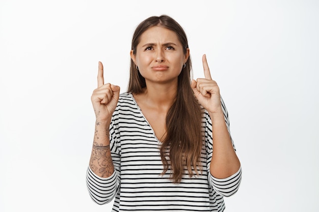 Disapleased girl grimacing from dislike, pointing fingers and looking up at smth displeasing, upsetting news, white background