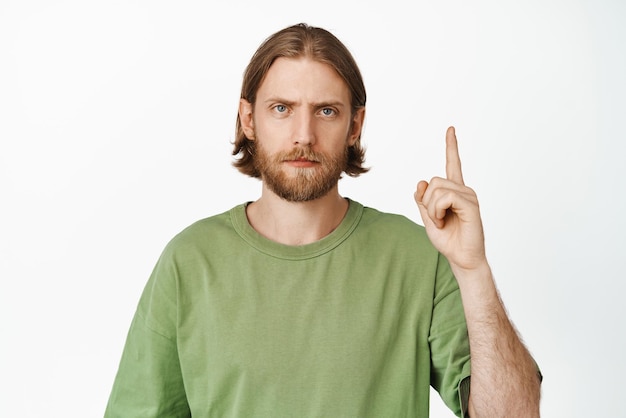 Disappointed adult bearded guy frowning upset or bothered pointing finger up showing advertisement on top fed up with smth standing against white background