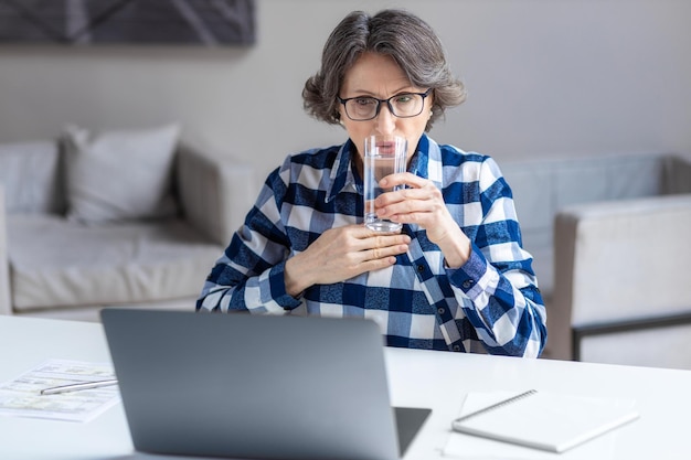 Disappointed aged woman sitting at the table feel frustrated by unexpected bad news on laptop from bank or cyberbullying concept