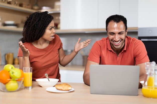 Disappointed angry young african american wife screaming on smiling husband guy play at laptop