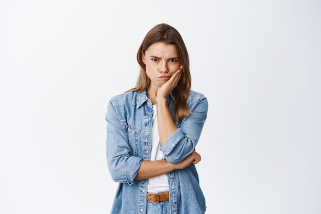 Disappointed and angry young woman frowning sulking and staring at camera annoyed feel jealous or displeased holding hand on cheek white backround