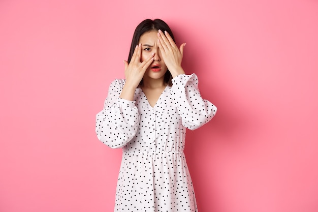 Disappointed asian woman peeking through fingers, open eyes and frowning displeased, staring with disdain at camera, standing over pink background.