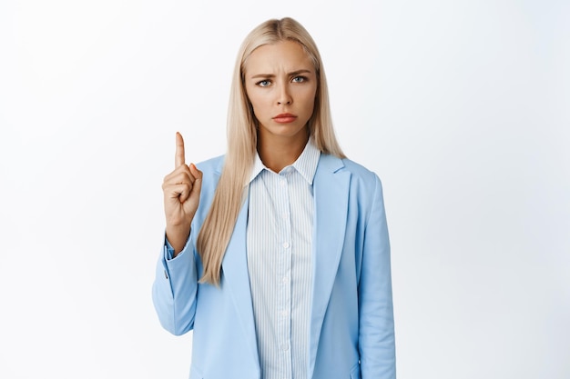 Disappointed corporate woman pointing finger up looking upset and frowning standing in suit over white background