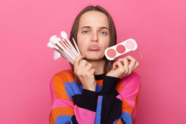 Photo disappointed displeased woman wearing sweater holding cosmetic brush powder looking with pout lips being in bad mood posing isolated on pink background