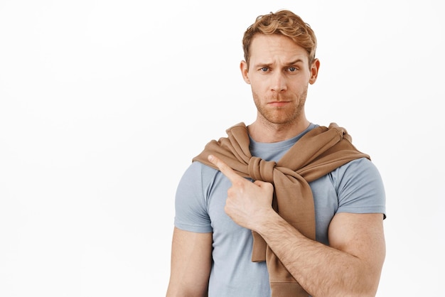 Disappointed and doubtful man with red hair and strong arms frowning looking with disbelief while pointing finger at upper left corner showing bad unimpressive thing white background