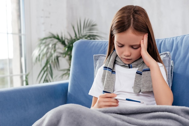 Disappointed girl wearing scarf and holding thermometer in hand while touching her head