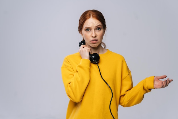 Disappointed sad young woman in stylish yellow sweater talking on retro phone and looking at camera